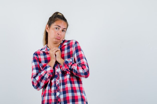 Young lady clasping hands on chest in checked shirt and looking helpless , front view.