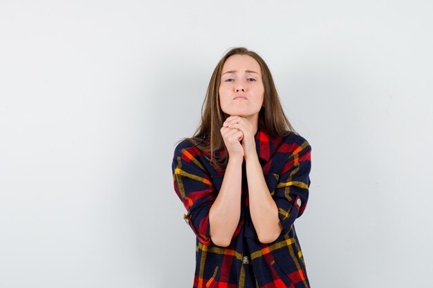 Young lady clasping hands in praying gesture in casual shirt and looking hopeful. front view.