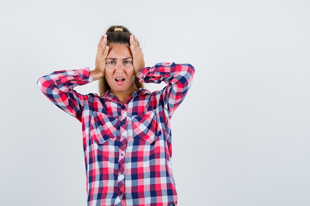 Young lady clasping head with hands in checked shirt and looking wistful , front view.