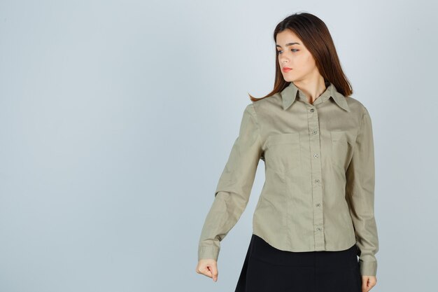 Young lady clenching fist with anger while looking down in shirt, skirt and looking offended , front view.