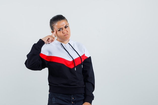 Young lady in colorful sweatshirt pointing at her head and looking angry , front view.