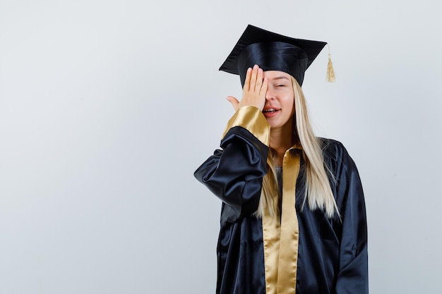Young lady covering eye with hand in academic dress and looking cute.