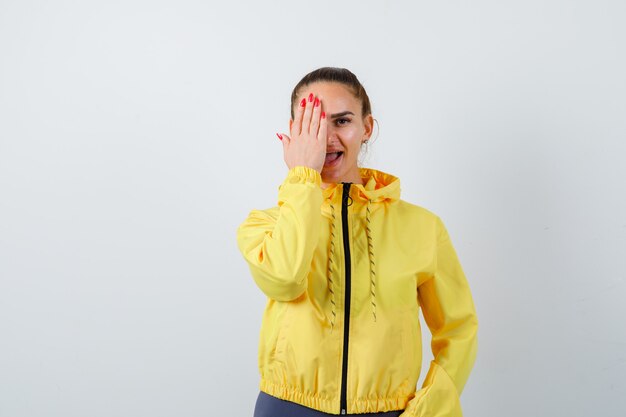 Young lady covering eye with hand in yellow jacket and looking positive , front view.