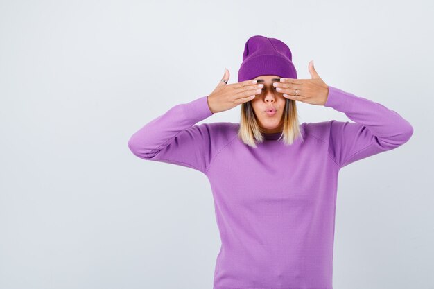 Foto gratuita giovane donna che copre gli occhi con le mani in maglione viola, berretto e sembra divertita, vista frontale.