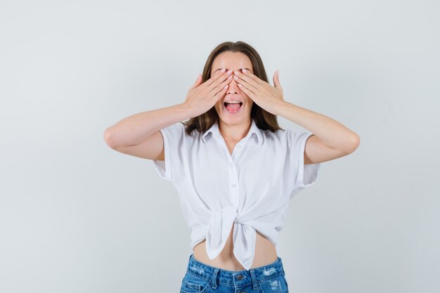 Young lady covering eyes with hands in white blouse and looking shy. front view.