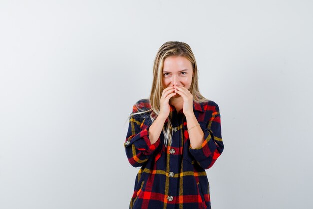 Young lady covering mouth with fingers in checked shirt and looking cute. front view.