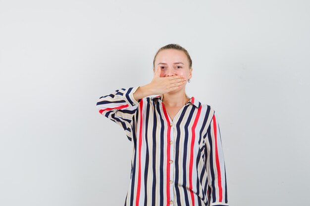 Young lady covering mouth with hand in striped shirt and looking positive , front view.