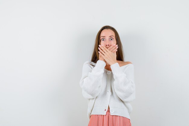 Young lady covering mouth with hands in cardigan and skirt looking puzzled isolated