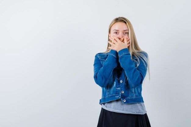 Young lady covering mouth with hands in t-shirt, denim jacket, skirt and looking confident.