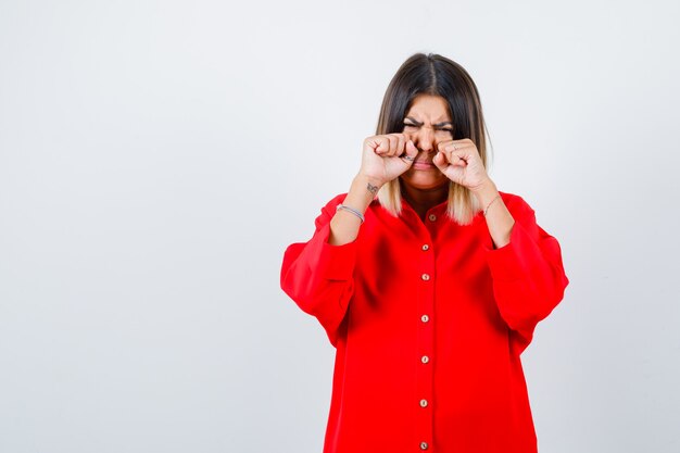 Young lady crying while rubbing eyes with hands in red oversize shirt and looking sorrowful , front view.