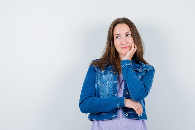 Young lady in denim jacket leaning cheek on hand, looking away and looking thoughtful , front view.