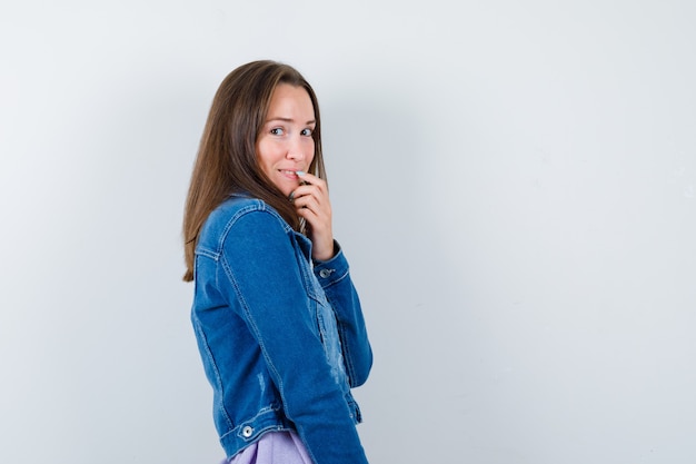 Young lady in denim jacket touching lip with finger and looking ashamed .