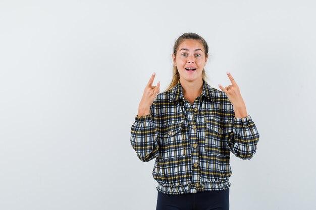 Young lady doing rock symbol in shirt, shorts and looking confident. front view.