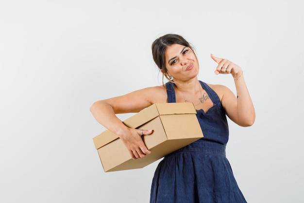 Young lady in dress holding cardboard box, pointing to the front and looking merry