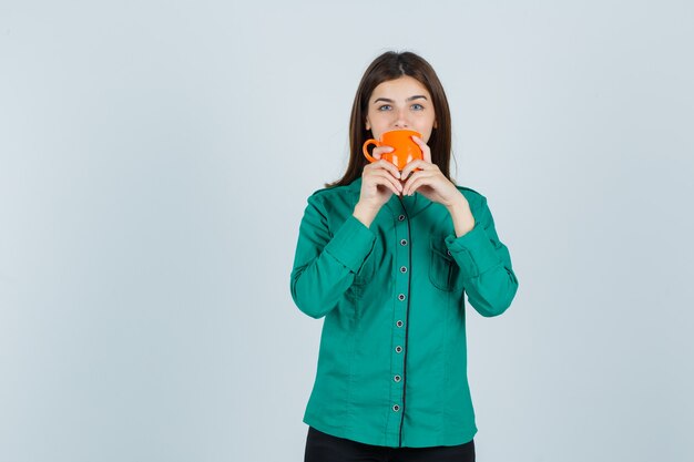 Young lady drinking tea from orange cup in shirt and looking confident. front view.