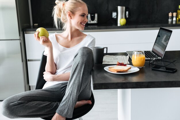 Young lady eating apple and using laptop in kitchen