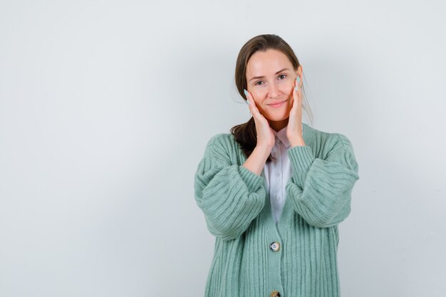 Young lady examining face skin by touching cheeks in blouse, cardigan and looking delicate , front view.