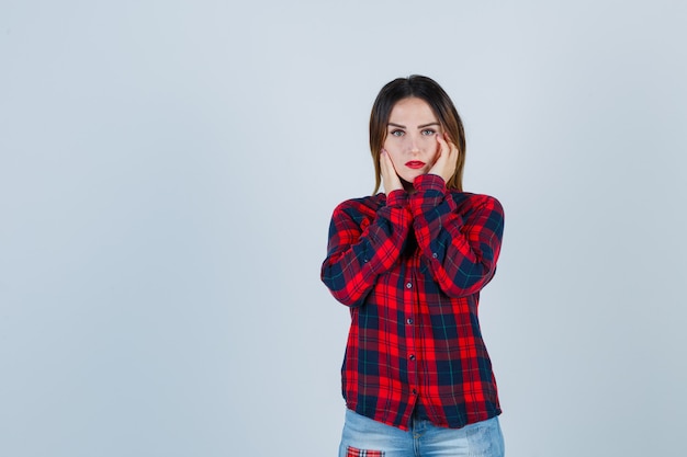 Young lady examining face skin by touching cheeks in checked shirt and looking delicate , front view.