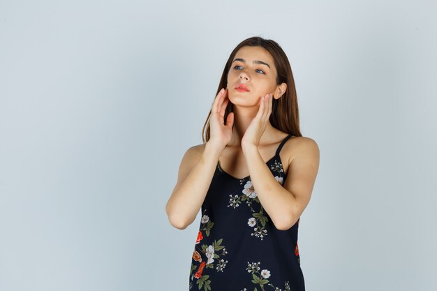 Young lady examining her face skin with hands in blouse and looking pensive , front view.