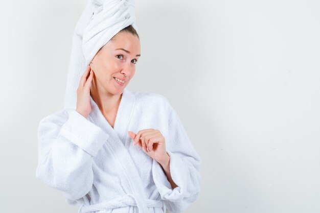 Young lady examining skin by touching her neck in white bathrobe, towel and looking careful. front view.