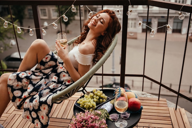 Young lady in floral skirt holds champagne glass and poses on terrace