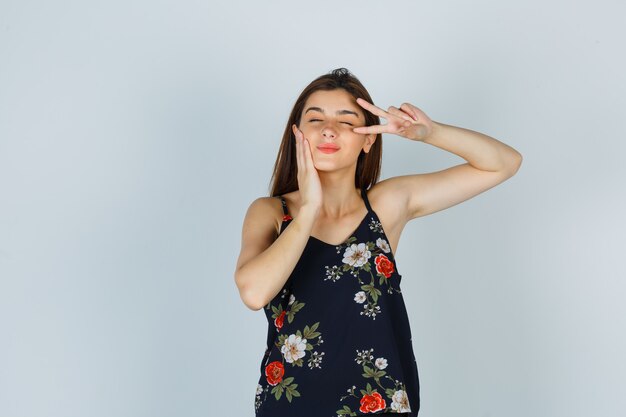 Young lady in floral top showing V-sign, leaning cheek on palm and looking happy