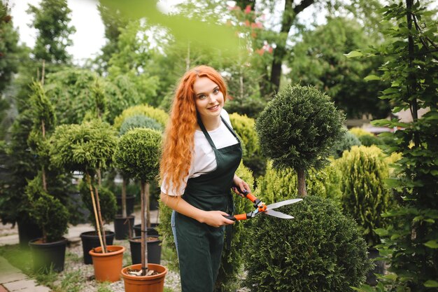 Young lady gardener with redhead curly hair standing in apron and holding big garden scissors while joyfully looking in camera outdoors