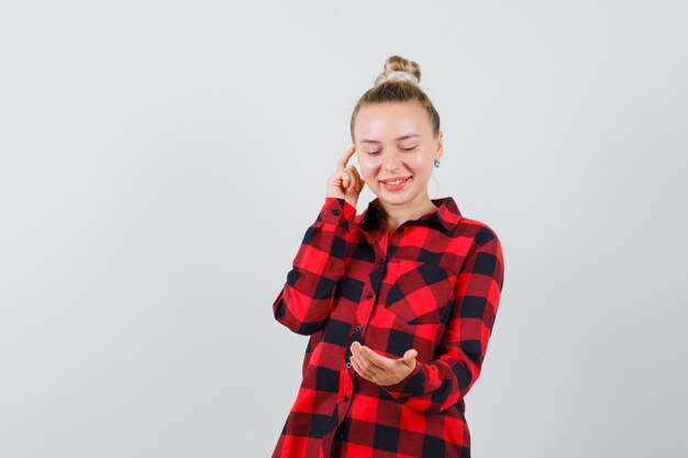 Young lady gesturing like looking at mobile phone in checked shirt and looking merry , front view.