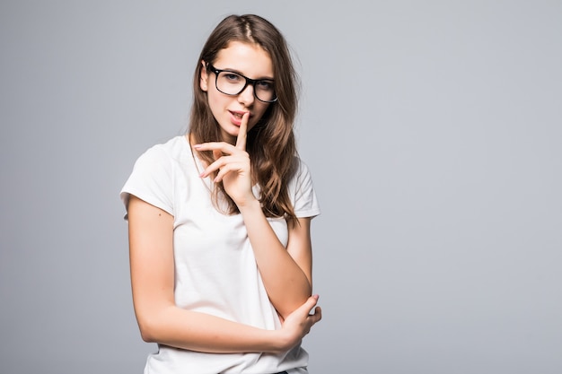 Young lady in glasses in white t-shirt and blue jeans shows shut up sign in front of white studio background