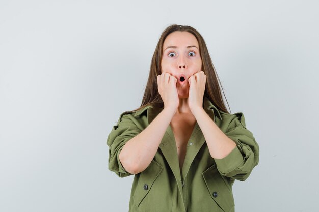 Young lady in green jacket holding fists on cheeks and looking surprised  