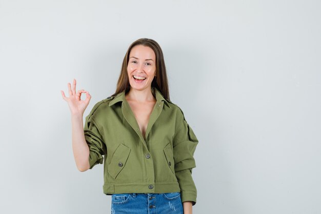 Young lady in green jacket, shorts showing ok gesture and looking jovial , front view.