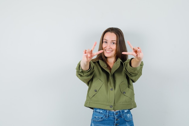 Giovane donna in giacca verde, pantaloncini che mostrano v-sign e che sembra vivace, vista frontale.