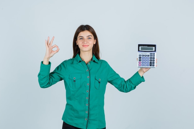 Молодая дама в зеленой рубашке держит калькулятор, показывает хорошо жест и выглядит жизнерадостной, вид спереди.