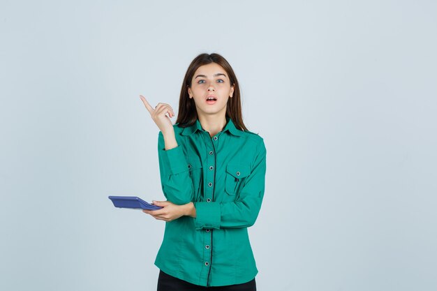 Молодая дама в зеленой рубашке держит калькулятор, показывая жест эврики, указывая вверх и выглядя удивленным, вид спереди.