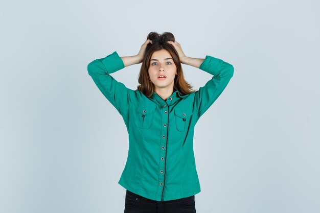 Young lady in green shirt holding hands on head and looking bewildered , front view.