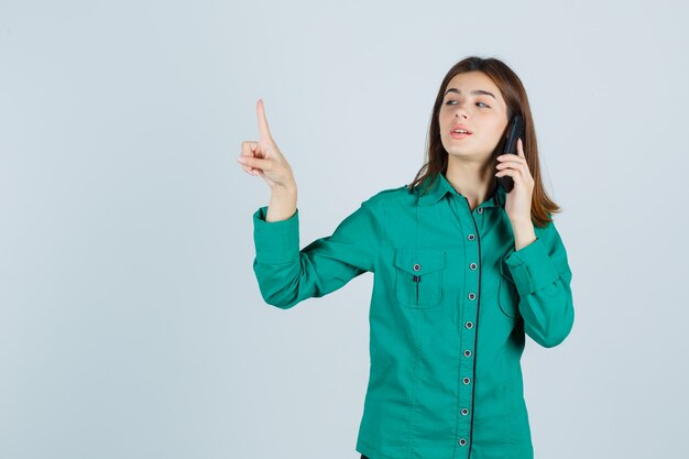 緑のシャツを着た若い女性が携帯電話で話し、細かいジェスチャーを保持し、自信を持って、正面図を示しています。