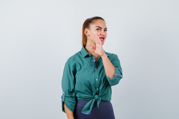 Young lady in green shirt telling secret behind hand and looking curious , front view.
