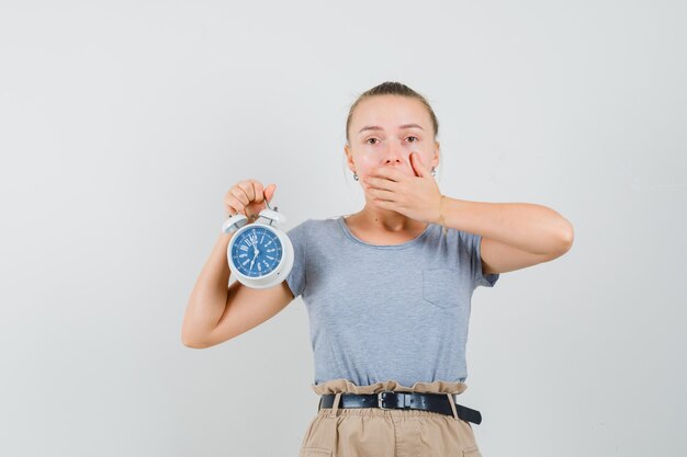 Tシャツ、ズボンで目覚まし時計を保持し、心配そうに見える若い女性。正面図。