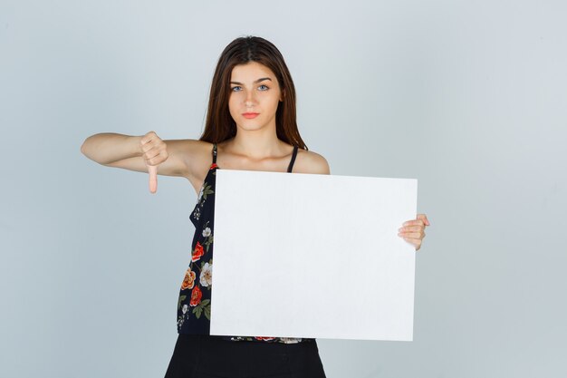 Young lady holding blank canvas, showing thumb down in blouse, skirt and looking displeased. front view.