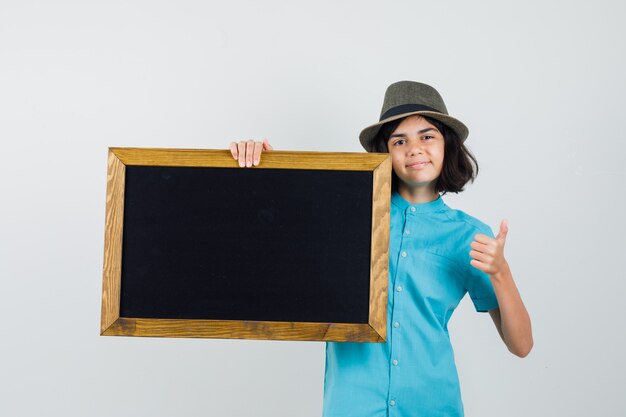 Молодая дама держит пустую рамку, показывая большой палец вверх в голубой рубашке, шляпе и выглядит оптимистично.