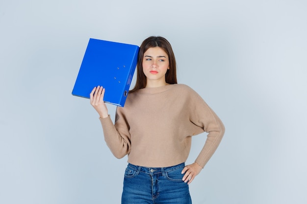 Young lady holding blue folder in beige sweater, jeans and looking disappointed. front view.