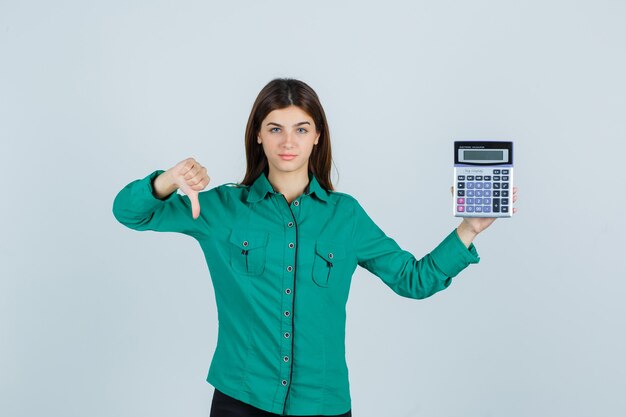 Young lady holding calculator, showing thumb down in green shirt and looking displeased. front view.