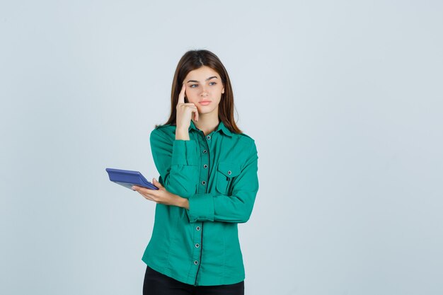 Young lady holding calculator while holding finger on temples in green shirt and looking pensive , front view.