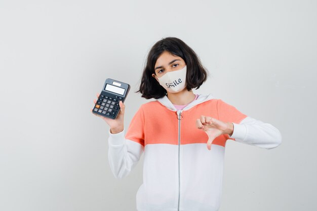 Young lady holding calculator while showing thumb down in hoodie, face mask and looking displeased.