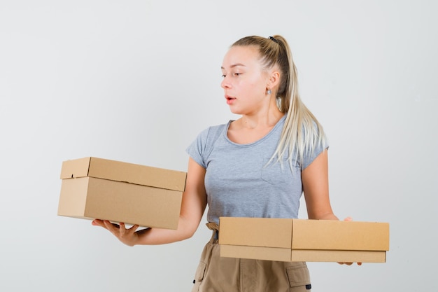 Young lady holding cardboard boxes in t-shirt, pants and looking focused , front view.