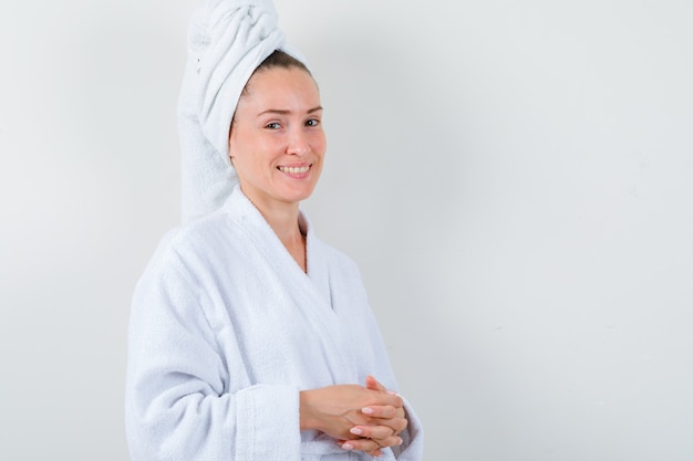 Young lady holding clasped fingers in front of her in white bathrobe, towel and looking cheery , front view.