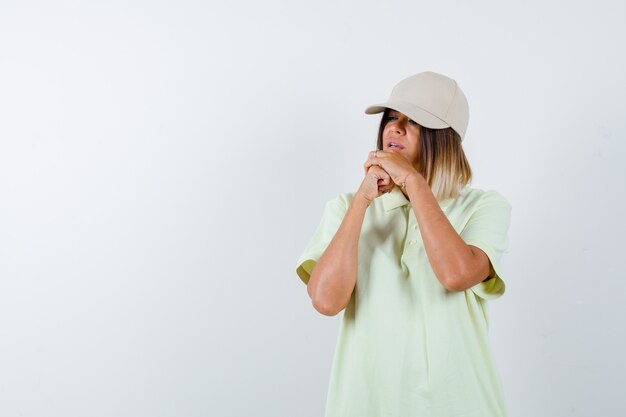 Young lady holding clasped hands near face in t-shirt, cap and looking hopeful , front view.