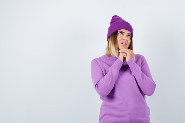 Free photo young lady holding clasped hands in praying gesture in purple sweater, beanie and looking hopeful , front view.