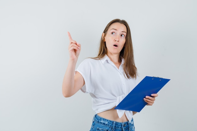 Young lady holding clipboard while pointing away in white blouse , front view.