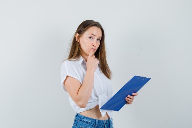 Young lady holding clipboard in white blouse and looking thoughtful , front view.